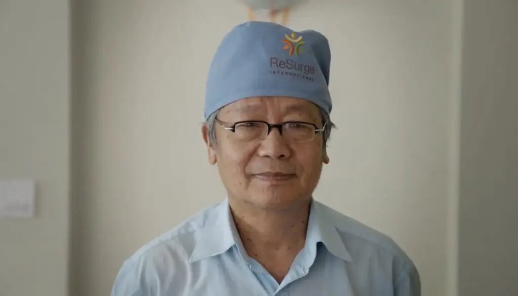 An older man, Dr. Shankar Man Rai, wearing glasses, a blue shirt, and a blue surgical cap with the ReSurge International logo stands indoors, facing the camera with a slight smile.