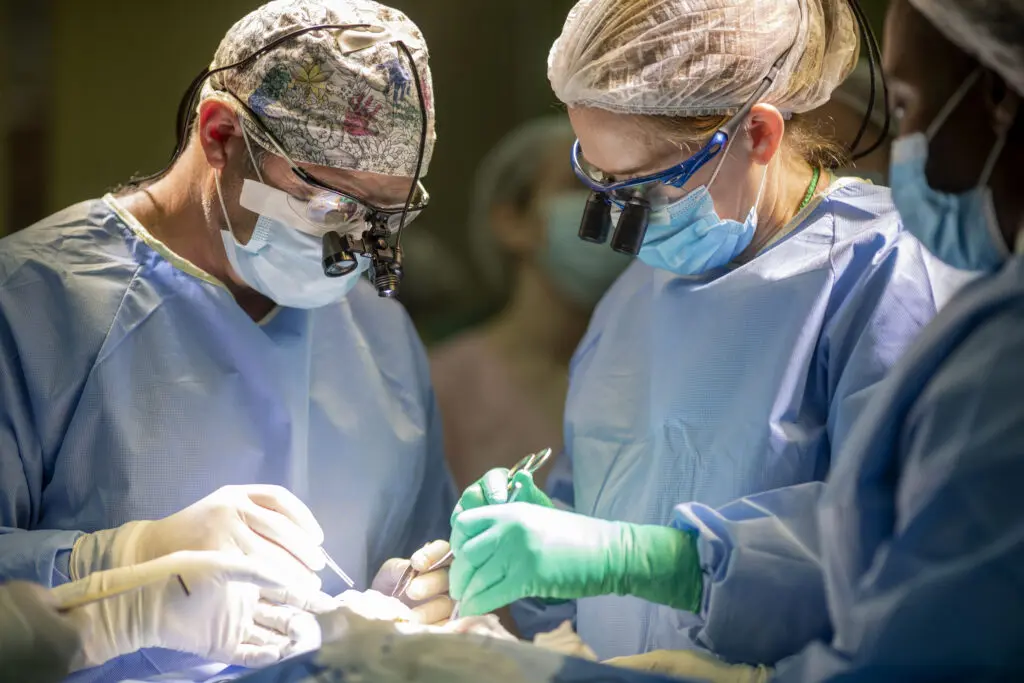 Three surgeons wearing scrubs, masks, surgical caps, and magnifying glasses focus intently while performing surgery under bright lights in an operating room.