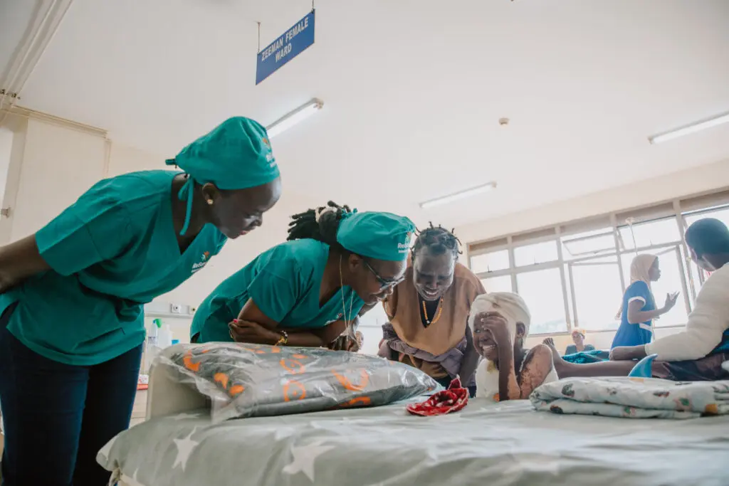 Three medical staff in teal scrubs smile and lean toward a laughing child lying in a hospital bed, while Razia, another adult, stands nearby. The scene is bright and cheerful, with large windows in the background.