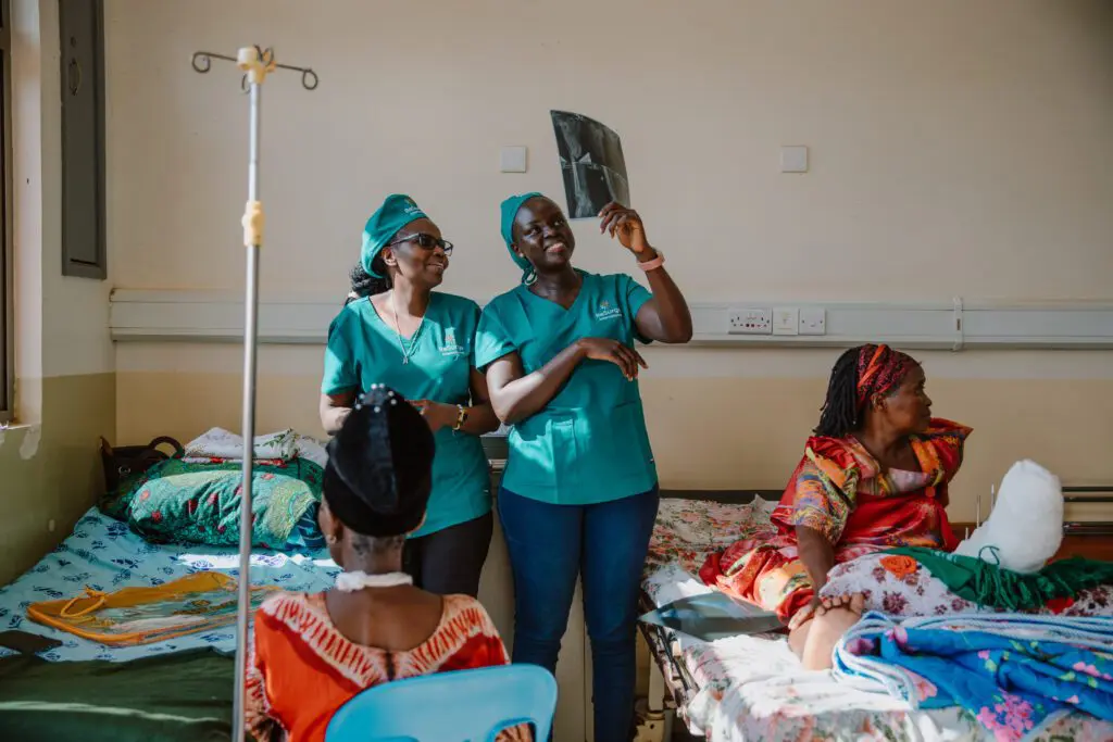 Two healthcare workers in green scrubs smile and examine an X-ray in a hospital ward, while two patients rest on beds nearby. The room is well-lit, and medical equipment is visible.