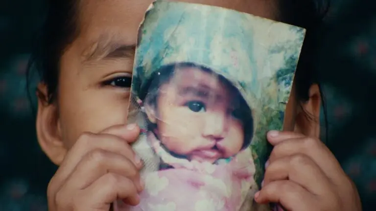 A child holds up a worn photo of a baby in front of their face, aligning the photo with their own features so that it appears as if the baby's face is part of their own.