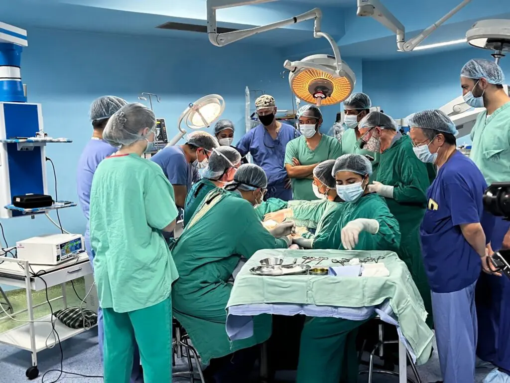 A group of medical professionals in scrubs and masks gathers around an operating table during surgery in a well-lit hospital operating room. Surgical instruments are laid out on a draped table.