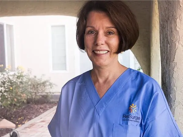 A woman wearing blue medical scrubs with a "ReSurge International" logo smiles while standing outside a building, with a window and garden visible in the background.