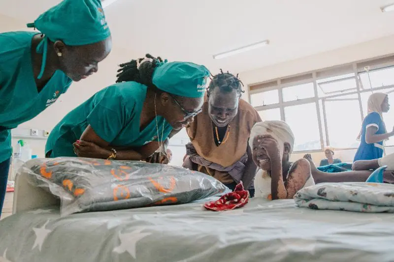Three women in medical uniforms and one in traditional clothing smile and lean toward a young girl with a bandaged head, who is smiling while sitting on a hospital bed covered with patterned blankets.