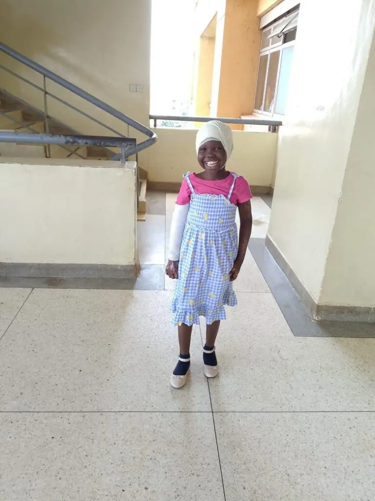 A smiling young girl with a bandaged arm stands indoors by a staircase, wearing a pink shirt, blue dress, dark socks, white shoes, and a white headwrap.
