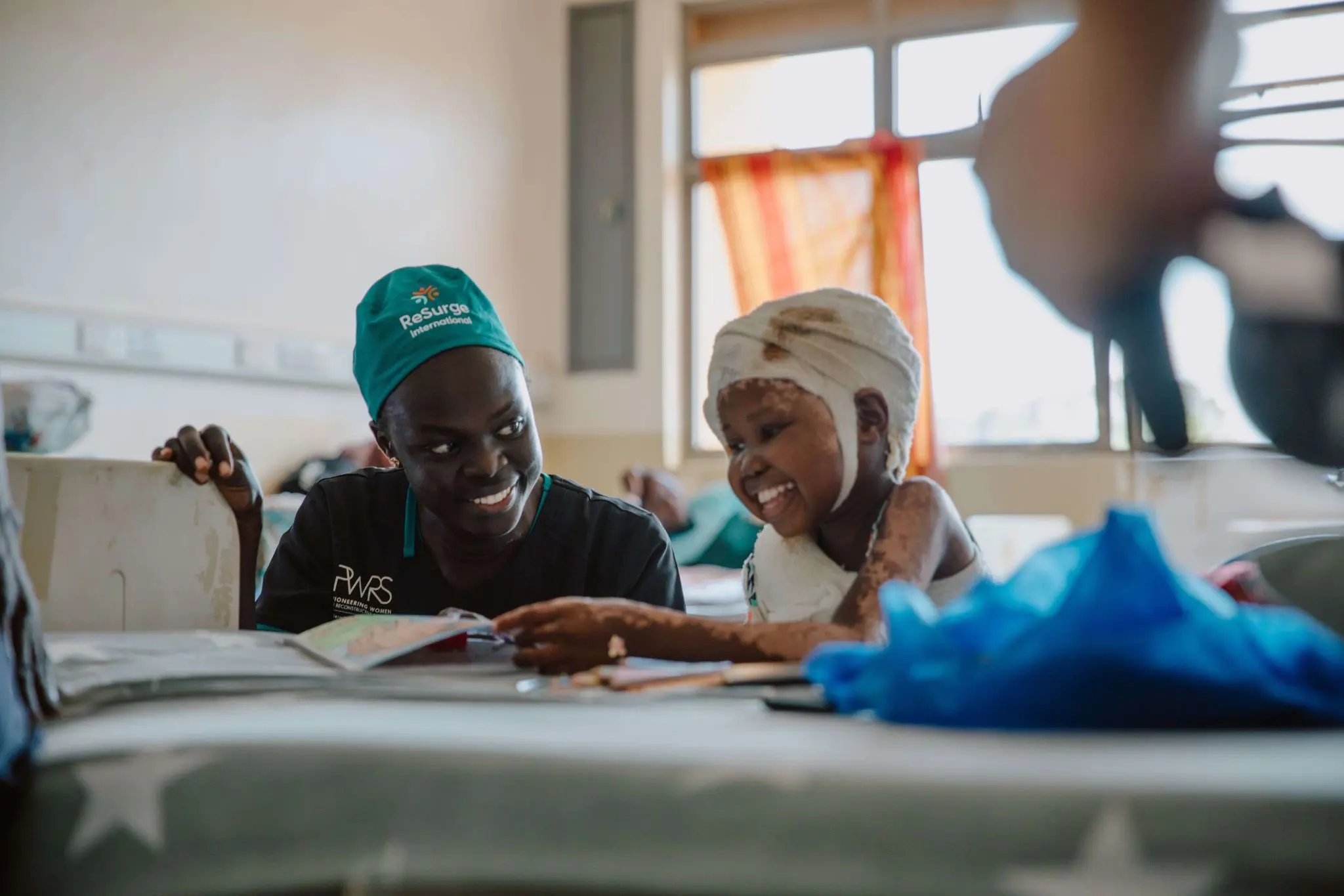 A smiling healthcare worker sits beside a young child with bandages on their head and arms, both engaged in an activity together in a brightly lit hospital room.