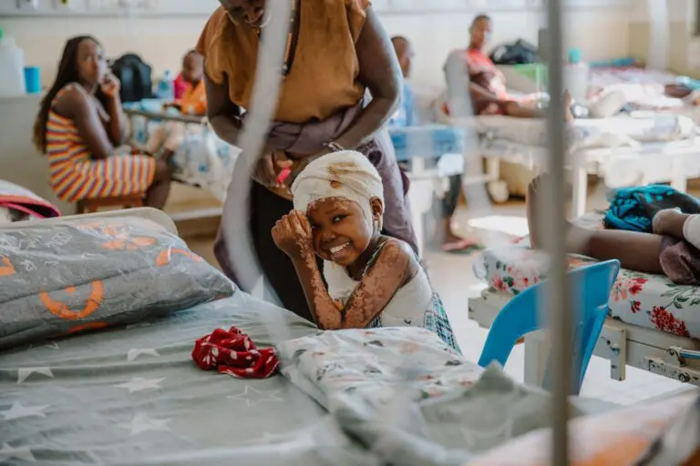 A young girl with bandages and visible scars smiles while sitting on a hospital bed. An adult stands nearby. Other patients and caregivers are visible in the background of the hospital ward.