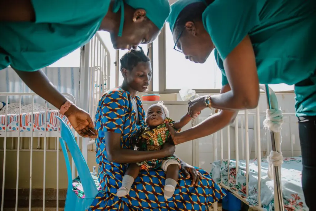 A woman holds a baby in her lap while two healthcare workers in green scrubs examine the child in a hospital room with cribs and natural light.