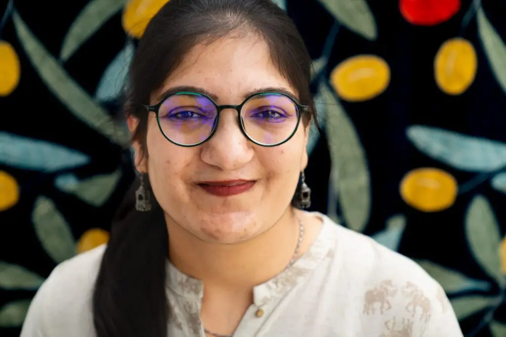A woman with glasses and earrings smiles in front of a dark background with colorful yellow and red patterns resembling fruits and leaves.