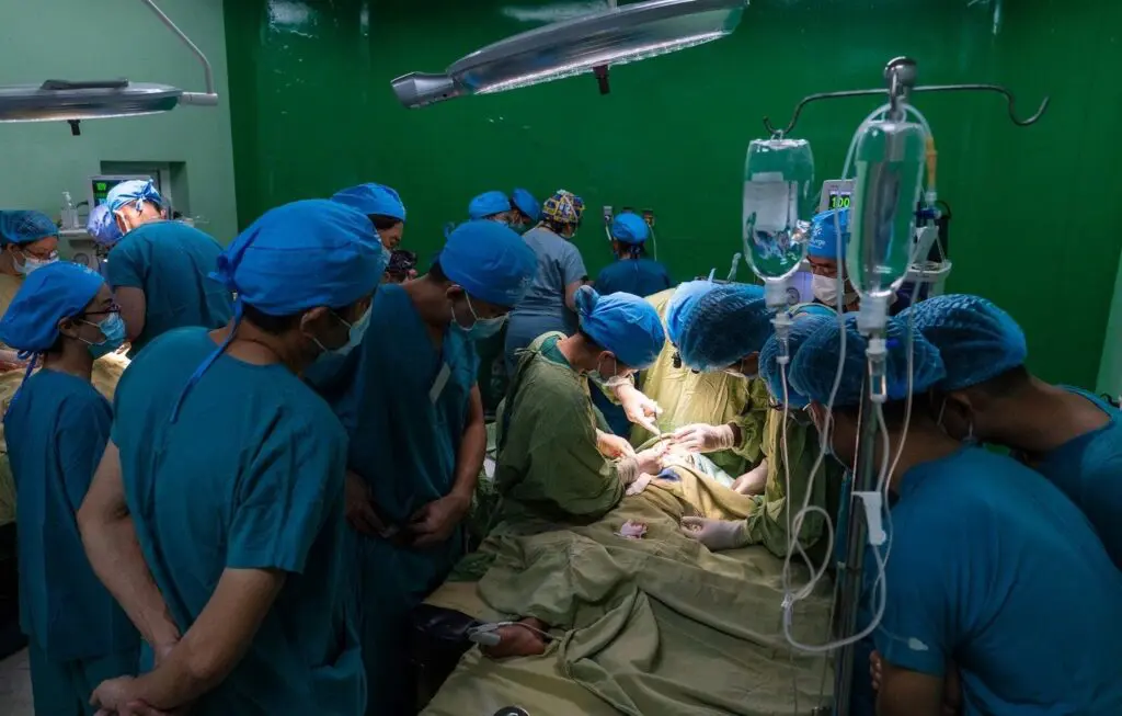 A team of surgeons and medical staff, wearing blue scrubs and masks, perform surgery in a brightly lit operating room with green walls. Medical equipment and IV stands are visible.