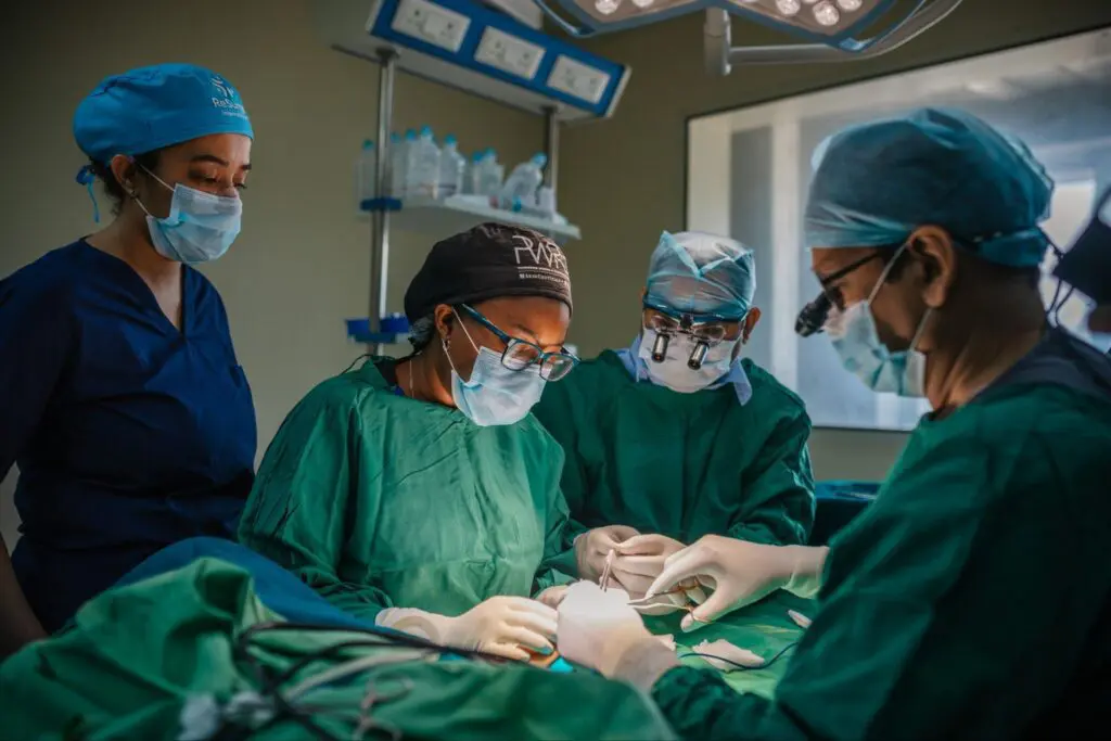 Four surgeons in scrubs and masks perform surgery in a well-lit operating room, focusing on the procedure. One assistant stands by, while medical equipment and lights are visible in the background.
