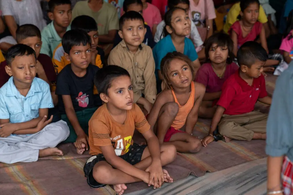 A group of children sit closely together on mats, listening attentively to someone off camera. Most look focused or curious, with some children wearing serious expressions.