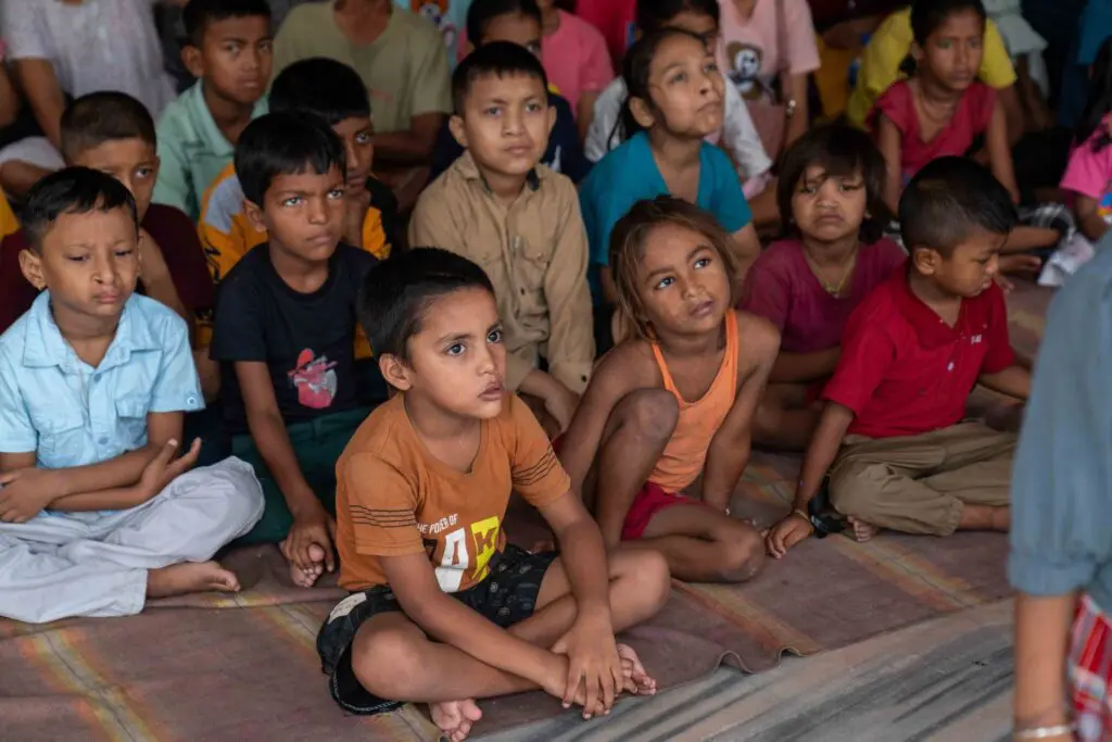 A group of young children sit cross-legged on a mat, attentively watching something out of frame. They wear casual clothing and appear focused and curious.