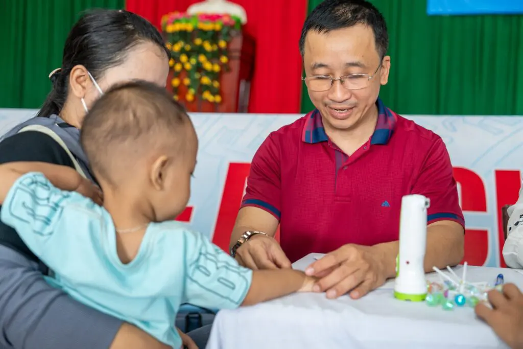 A man in a red shirt examines a young child’s hand at a medical table, while a woman holds the child. Medical supplies and colorful backgrounds are visible.