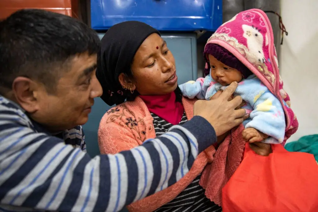 A man and woman, both in warm clothing, lovingly interact with a baby dressed in a colorful, hooded outfit. The woman holds the baby while the man gently touches the baby's chin.