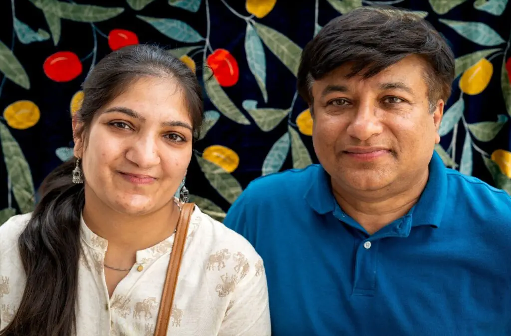 Two people smiling in front of a dark blue background with colorful leaves and fruit patterns. The person on the left has long dark hair and wears a light shirt; the person on the right wears a blue collared shirt.