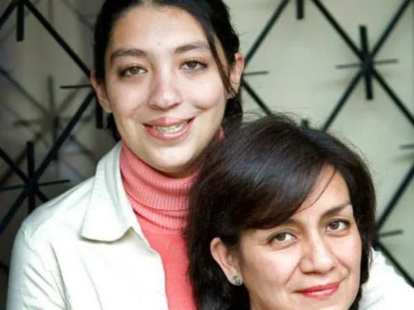 Two women pose closely together and smile at the camera. One stands behind the other, wearing a white jacket and pink turtleneck. The woman in front has dark hair and wears a light-colored top. A patterned background is visible.