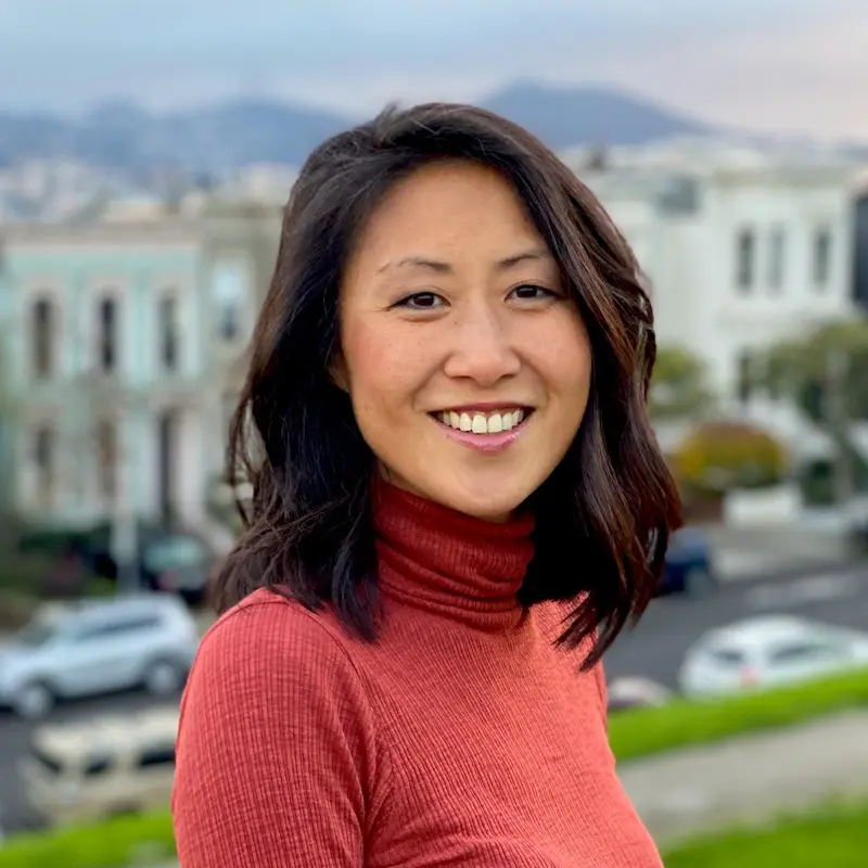 A woman with shoulder-length dark hair, wearing a rust-colored turtleneck, smiles outdoors with blurred houses, cars, and hills in the background.