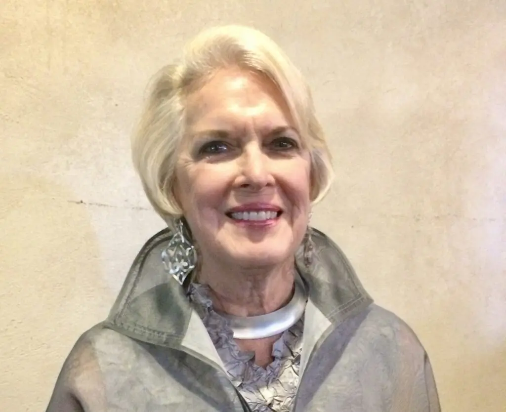 An older woman with short blonde hair smiles at the camera, wearing a gray, elegant outfit and large silver earrings, standing against a beige textured wall. The poised figure evokes the timeless charm of Kathryn E. Johnson.