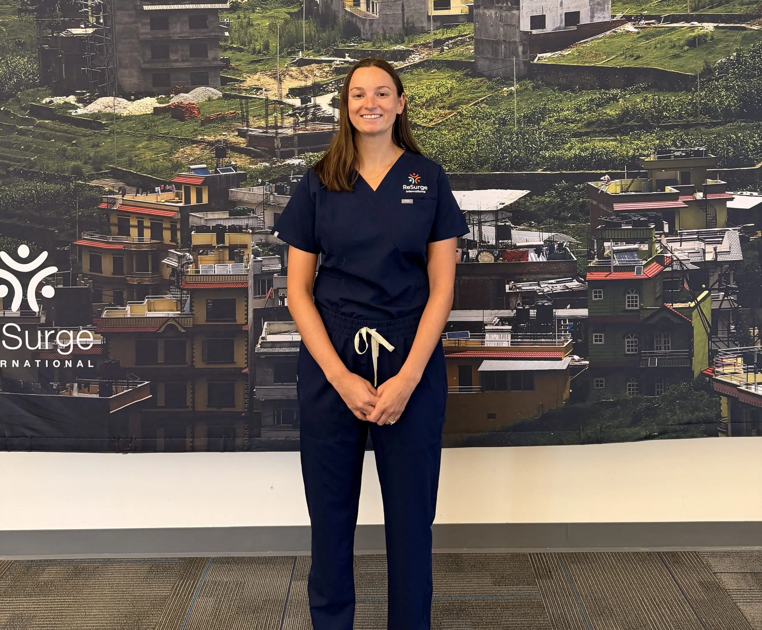 A woman in navy blue medical scrubs stands smiling in front of a large mural showing a densely packed cityscape with colorful buildings. The Surge International logo appears on the wall to her left, identifying her as Dr. Kelsi Krakauer, the 2026 ReSurge Laub Fellow.