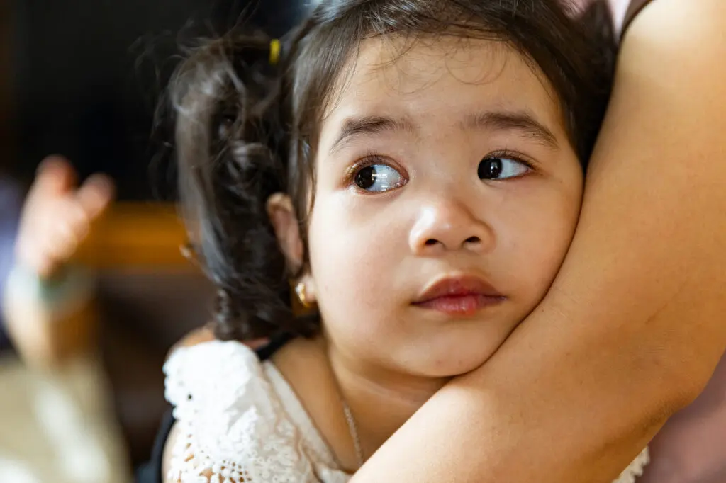 A young Ha Nhi girl with dark hair and ptosis looks to the side while being held closely by an adult, her expression appears anxious or worried, perhaps due to concerns about her sight.