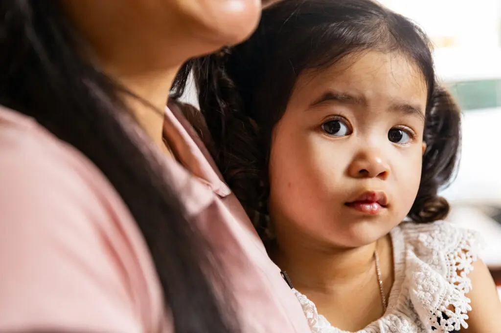Young Ha Nhi girl with wavy hair and white lace top sits closely beside an adult, looking slightly to the side with a calm, thoughtful expression, her gaze hinting at both ptosis and a keen sense of sight.