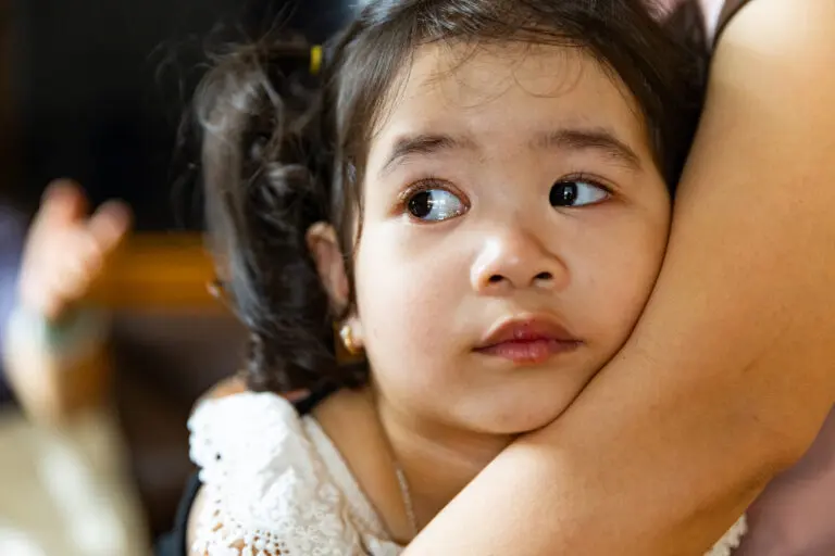 A young Ha Nhi girl with dark hair and ptosis looks to the side while being held closely by an adult, her expression appears anxious or worried, perhaps due to concerns about her sight.