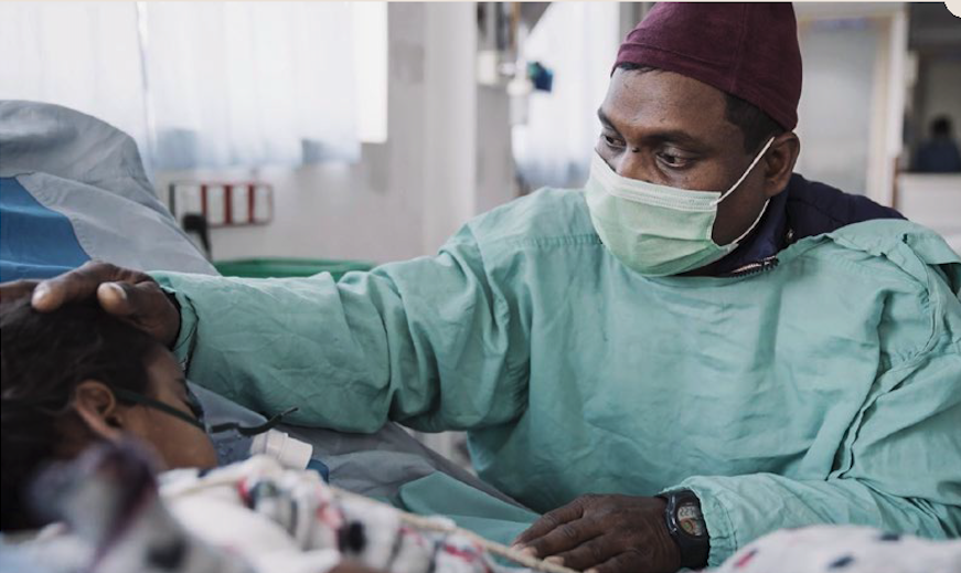 A medical professional in scrubs and a mask gently touches the head of a patient lying in a hospital bed, providing comfort and care as part of their dedicated reconstructive surgical care.