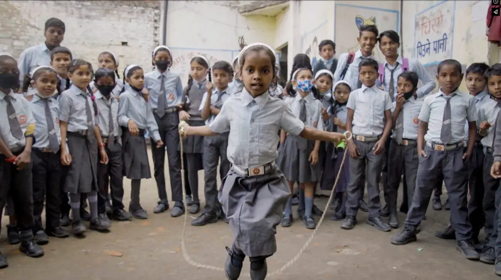 A young schoolgirl in uniform jumps rope while classmates in similar uniforms watch and smile in a sunlit schoolyard—a joyful scene captured during the filming of a documentary film about reconstructive surgery in Nepal.