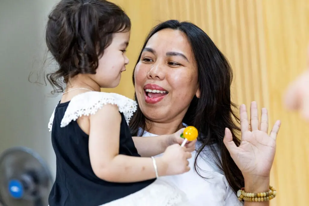 A smiling woman gives a high five to a young girl in a black and white dress holding a lollipop. They are indoors with a light-colored wall in the background.