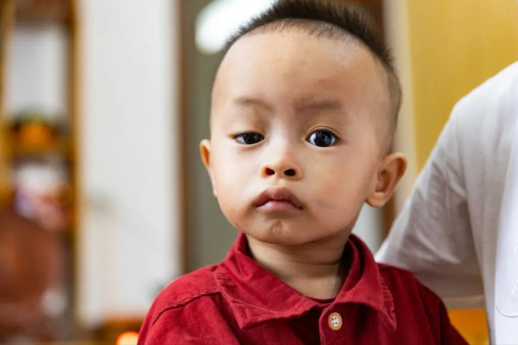 A young child wearing a red collared shirt looks at the camera with a neutral expression. The child is indoors, and an adult wearing a white shirt is partially visible beside them.