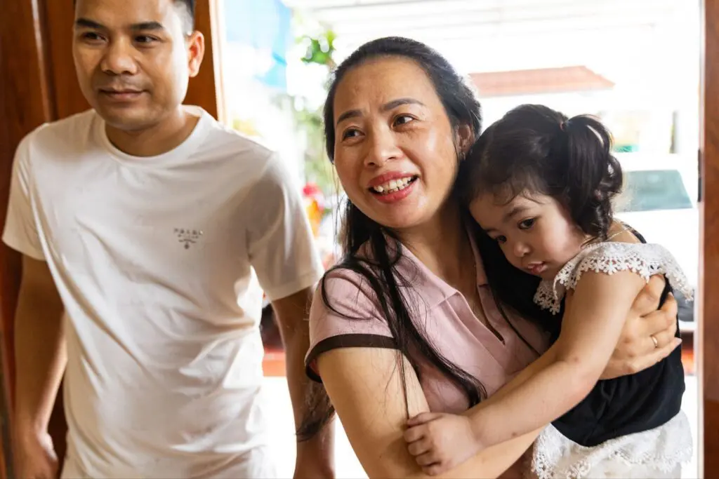 A smiling woman holds a young girl in her arms while a man in a white shirt stands nearby. They appear to be indoors, near an open door, with natural light coming in.