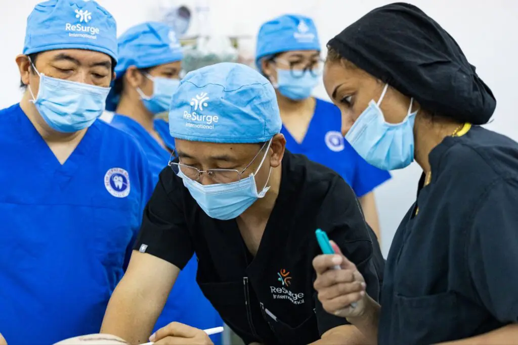 A group of surgeons wearing blue scrubs, blue caps, and face masks focus on a medical procedure in an operating room. One surgeon holds a pen and others observe closely.