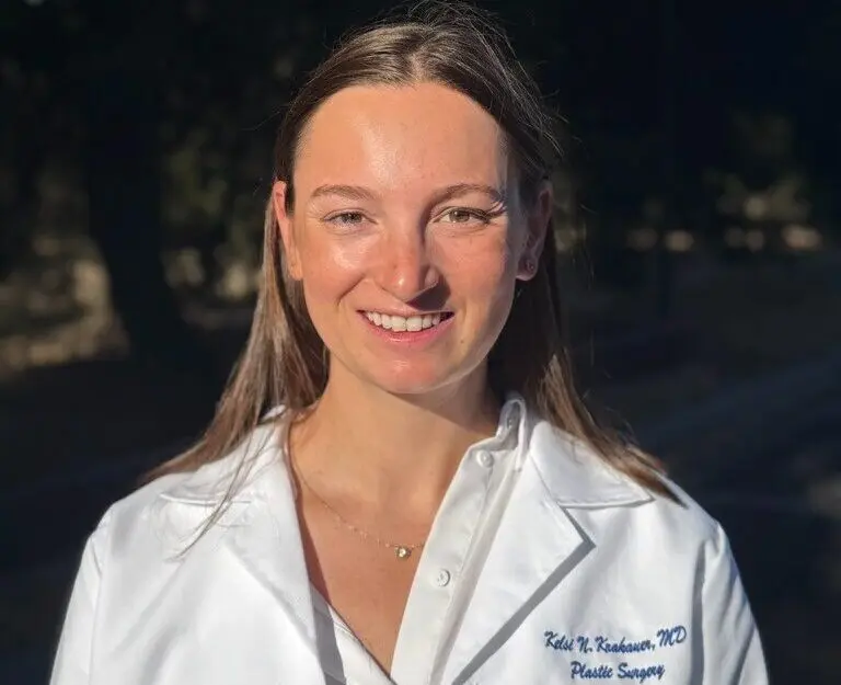 A woman in a white lab coat smiles outdoors in sunlight, with trees and a path in the background. The lab coat reads “Plastic Surgeon” and “MD” on the chest.