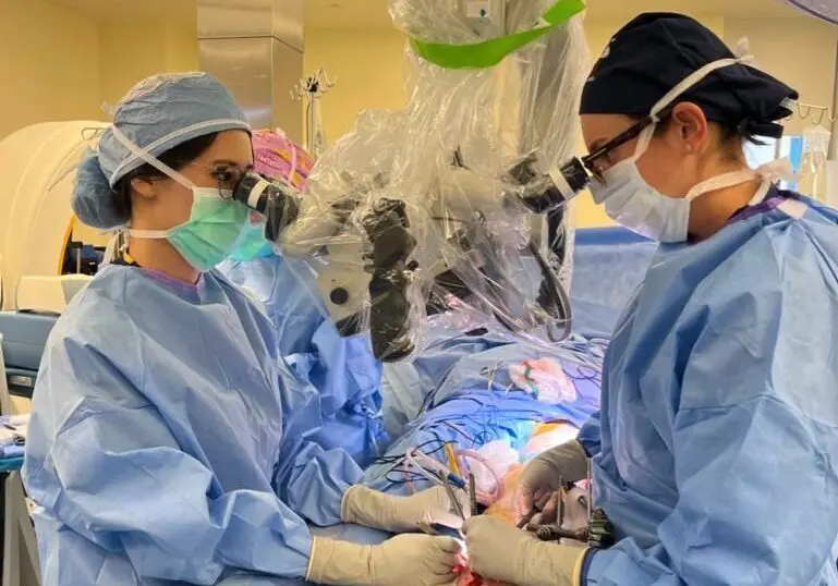 Two surgeons in blue scrubs and masks perform surgery in an operating room, using magnifying equipment. Surgical tools and gauze are on a tray in the foreground. Bright lights illuminate the sterile environment.