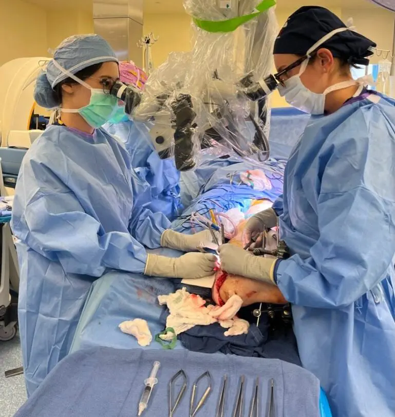 Two surgeons in blue scrubs and masks perform surgery in an operating room, using magnifying equipment. Surgical tools and gauze are on a tray in the foreground. Bright lights illuminate the sterile environment.