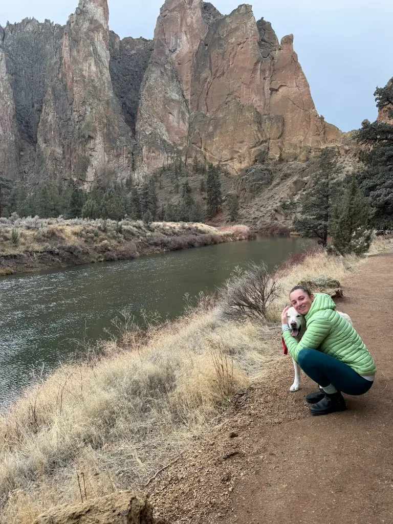 A person in a green jacket, possibly Dr. Kelsi Krakauer, ReSurge Laub Fellow 2026, crouches next to a white dog beside a river, with tall rocky cliffs and sparse trees in the background.