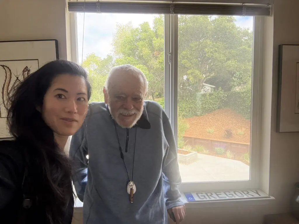 A woman and an older man stand smiling indoors by a window with a backyard view. Sunlight fills the room, creating a warm atmosphere that reflects their sense of care for one another. A sign with “Gregory” is visible on the windowsill.