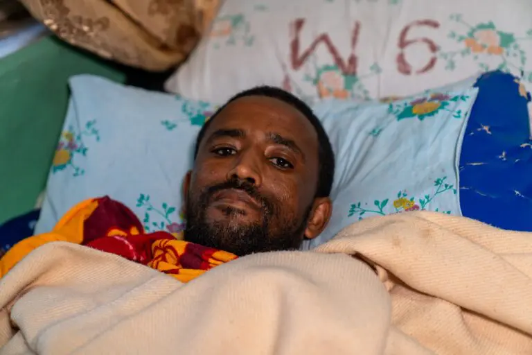 A man with a beard lies in bed, covered by a beige blanket. He looks at the camera, with floral-patterned pillows and a pillow marked “W6” behind his head.