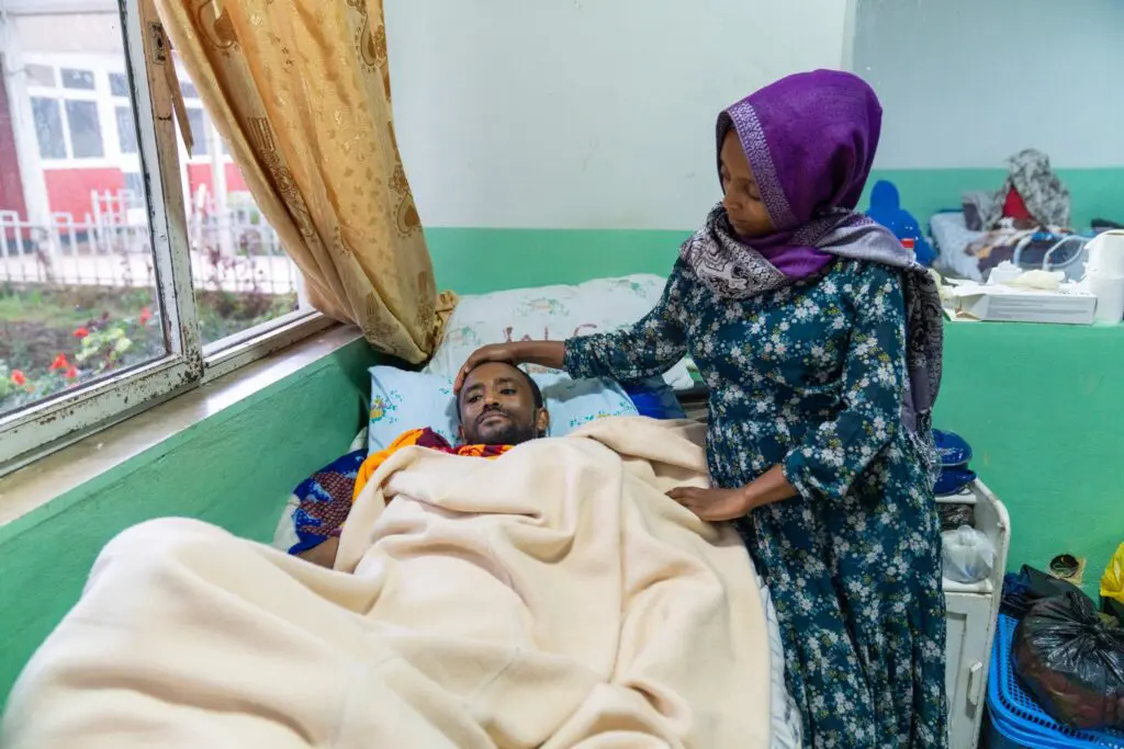 A woman in a patterned dress and headscarf stands by a hospital bed, gently touching the head of a man who is lying under a blanket near a window.