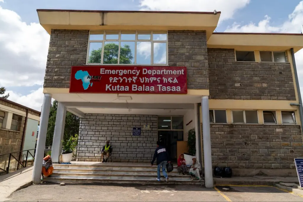 A hospital emergency department entrance in Ethiopia with a red sign in English and local script. Several people are sitting or standing near the doorway, perhaps awaiting care such as reconstructive surgery that recently saved a man’s leg. The stone building features large windows, welcoming patients and visitors alike.