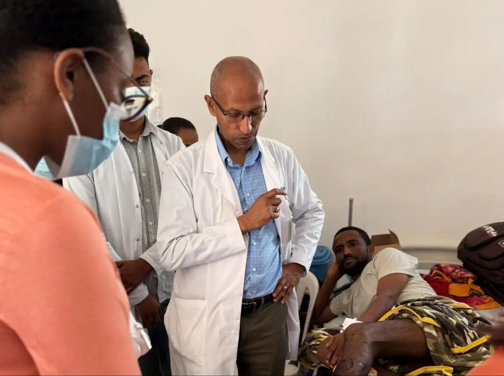 Doctors in white coats examine a patient lying on a hospital bed. One doctor appears thoughtful, while another is listening. The patient’s leg is bandaged following reconstructive surgery Ethiopia, and medical equipment is visible nearby.