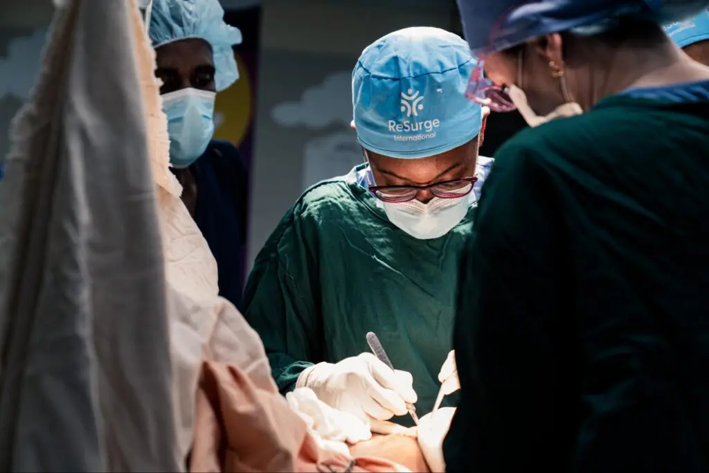 A surgeon in scrubs and a mask performs reconstructive surgery on a patient with a leg injury, assisted by other medical staff in surgical attire, in a well-lit operating room in Ethiopia.