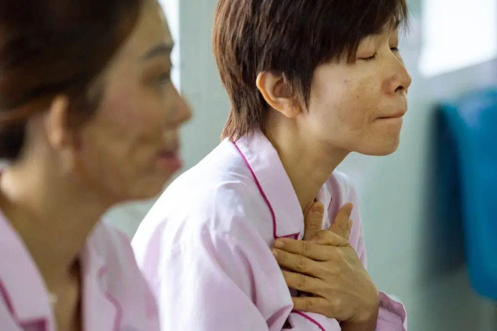 A woman in a light pink uniform sits with her eyes closed and her hands held against her chest, offering patient support to another woman slightly out of focus in the foreground.