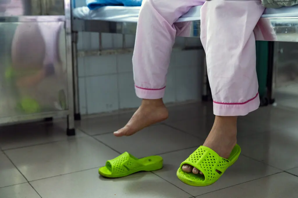 A patient recovering from breast reconstruction sits on a hospital bed, wearing pink pants with one foot in a bright green perforated slide and the other bare.