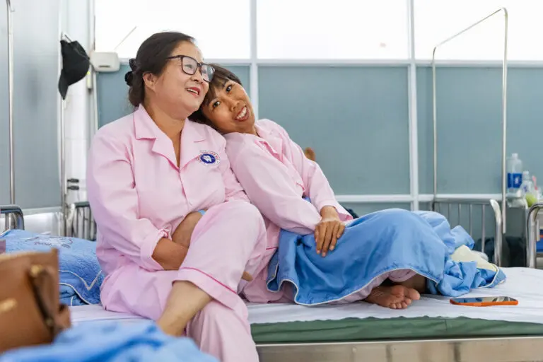 Two women in matching pink hospital pajamas sit side-by-side on a hospital bed, with one resting her head affectionately on the other’s shoulder. Both ReSurge patients and breast cancer survivors about to receive microsurgery
