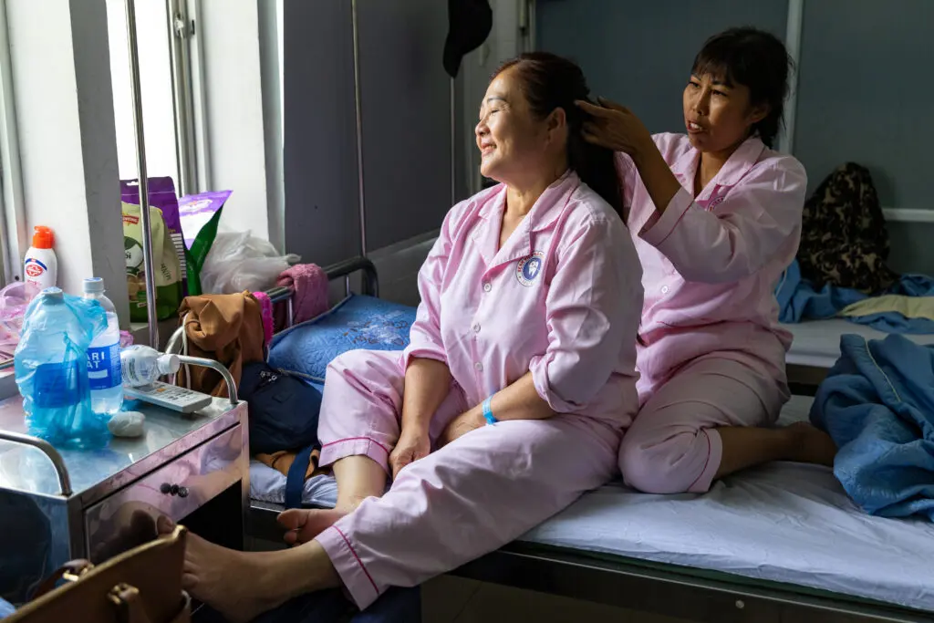 A woman in pink pajamas sits on a hospital bed recovering after breast reconstruction while another woman brushes her hair.