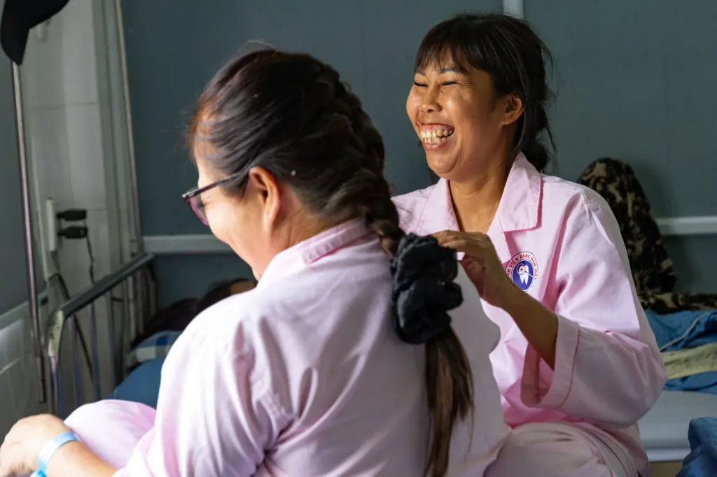 A woman in a pink hospital gown braids another patient’s hair as they both laugh, sharing a moment of recovery after breast reconstruction.
