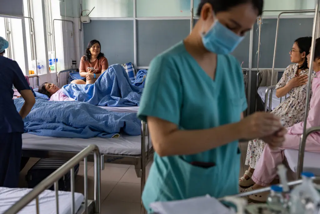 A medical worker in scrubs and a face mask in the foreground, with patients recovering from breast reconstruction after cancer and their visitors sitting on hospital beds in the background.