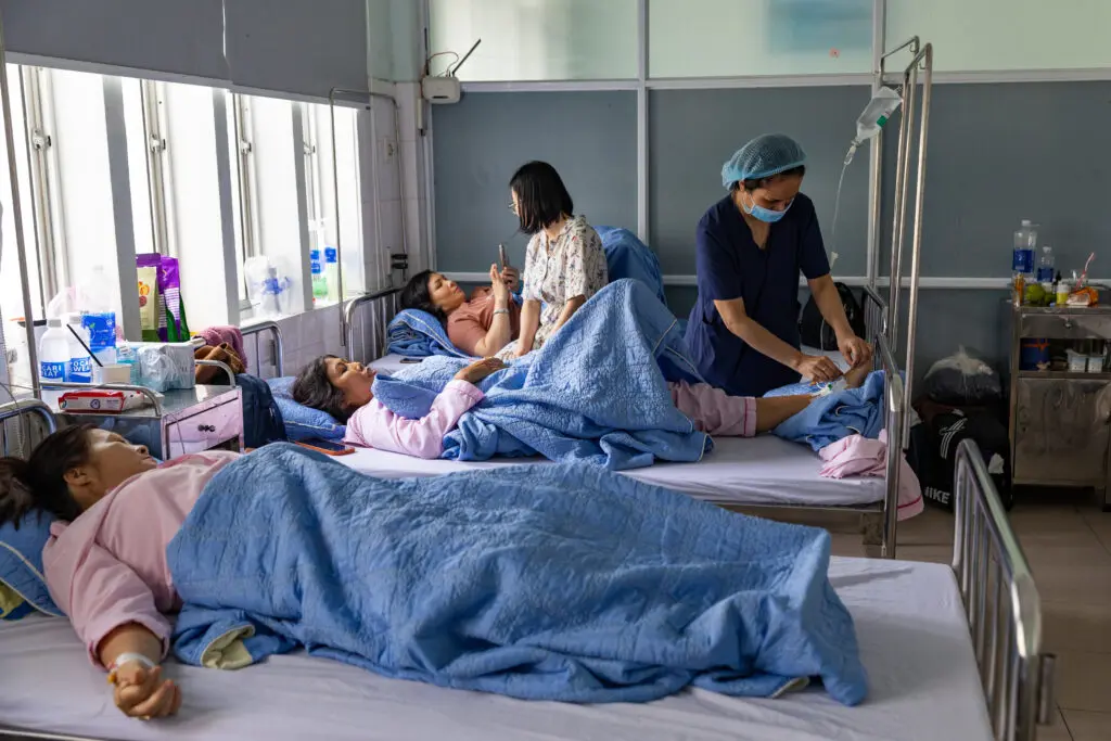 A nurse adjusts an IV line for a patient recovering from breast reconstruction after cancer in a shared hospital room, while a visitor sits nearby.